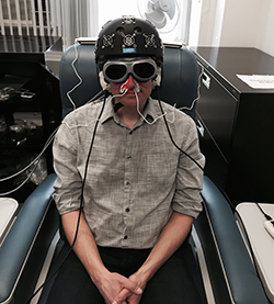 A man demonstrates equipment, including an LED helmet, intranasal diodes, LED cluster heads placed on the ears, and goggles.
