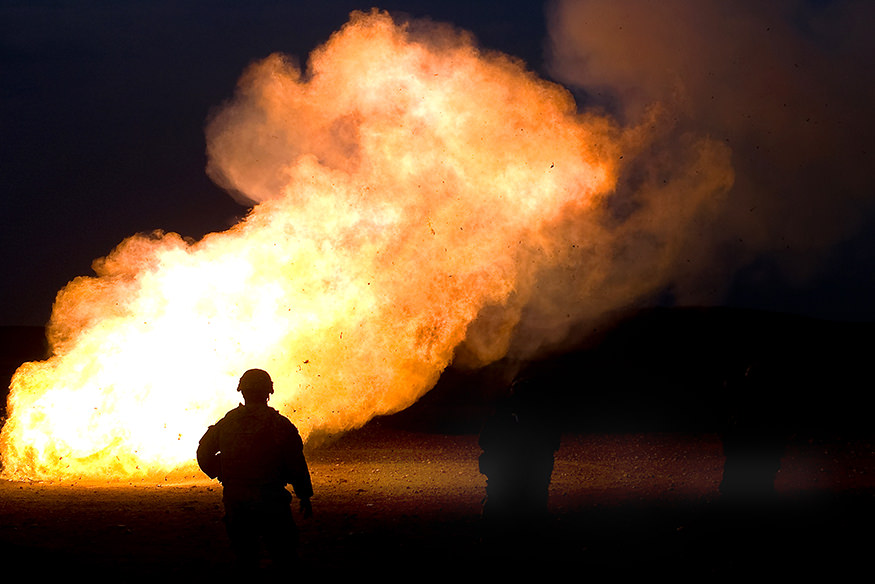 A soldier stands in silhouette against a large flame from the disposal of mortar charges.