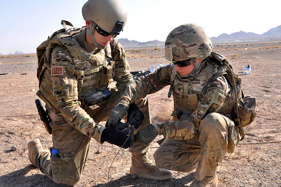 Two U.S. soldiers in full uniform tape a detonation cord during explosives training in a desert.